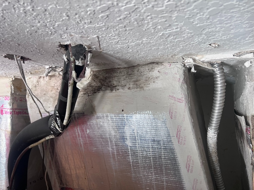 Close-up of black HVAC tape wrapped around a duct connection at a ceiling penetration, with dark mold staining visible on the insulation panel below