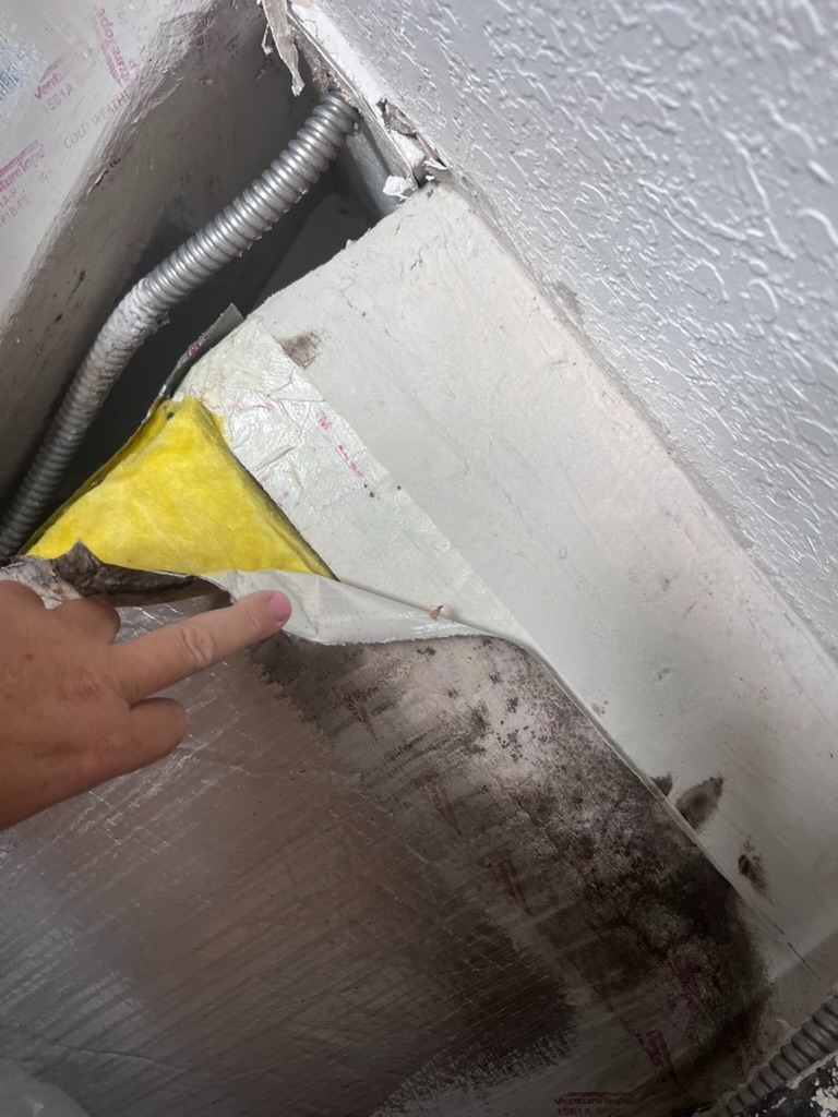 A hand pointing at a section of failed tape seal at a duct connection, with mold visible below the gap and yellow fiberglass insulation exposed